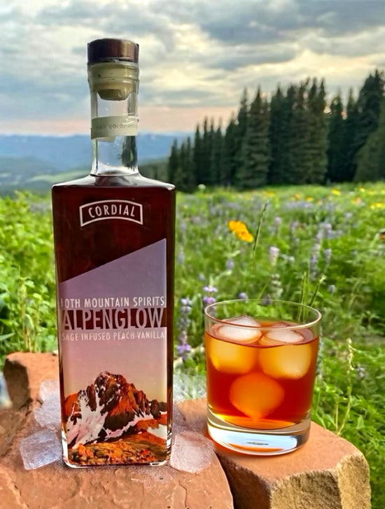 Bottle of Cordial Alpenglow with a glass of iced drink in a scenic outdoor setting.