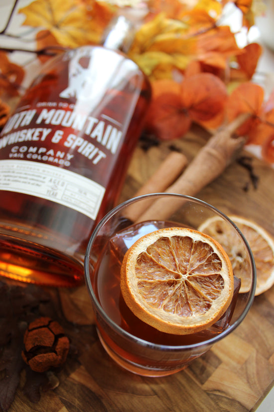 Bottle of 10th Mountain Whiskey & Spirit Company on a wooden surface with a glass of whiskey and dried orange slices.
