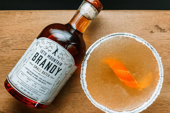 Bottle of 10th Mountain Brandy next to a cocktail with an orange twist on a wooden surface