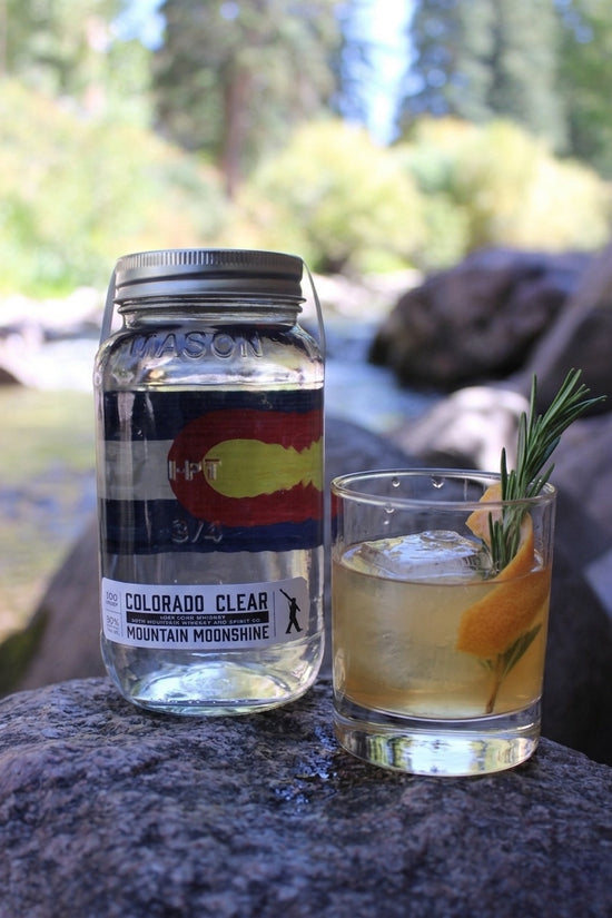 Bottle of Colorado Clear Mountain Moonshine and a cocktail with a garnish on a rock with a natural background.