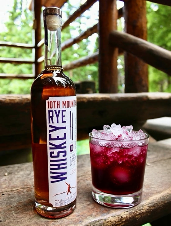 Bottle of 10th Mountain Rye Whiskey next to a glass of red cocktail with ice on a wooden surface outdoors.