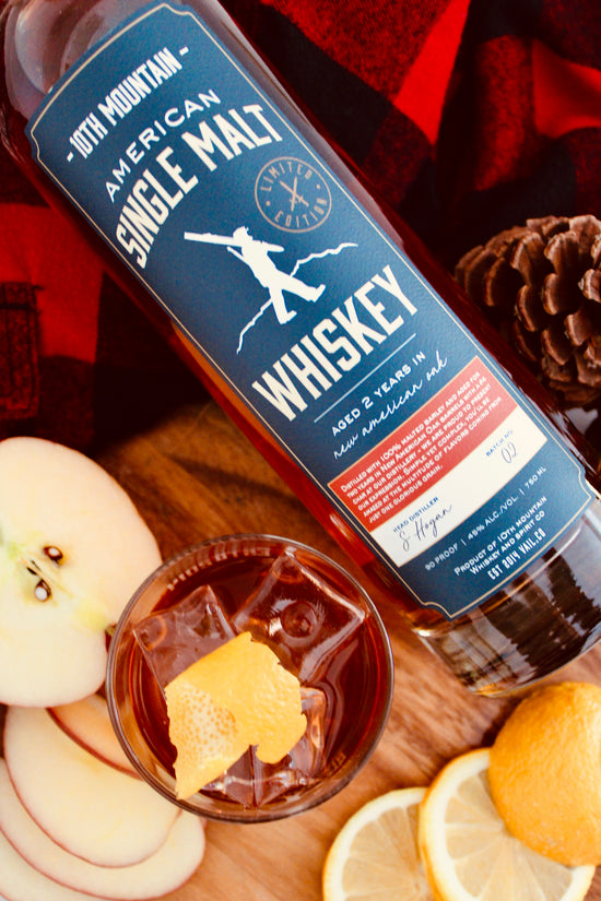 Bottle of 10th Mountain American Single Malt Whiskey with a glass of whiskey and fruit on a wooden surface.